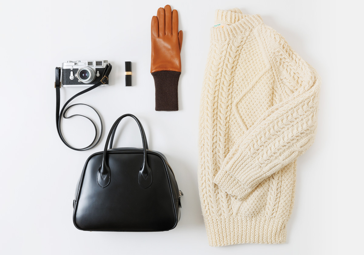 白い背景に、黒いハンドバッグ、カメラ、口紅、茶色いKURODAクロダの革手袋、白いニットが上品に配置されている、ファッションアイテムのフラットレイ写真